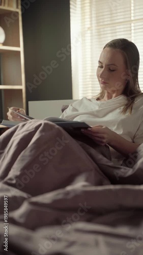 Relaxed young woman sitting comfortably in bed under a warm duvet, sketching with a pencil in her notebook while enjoying a quiet morning at home in a sunlit bedroom