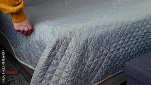 Person arranging bed linens, smoothing quilt on mattress in cozy bedroom setting