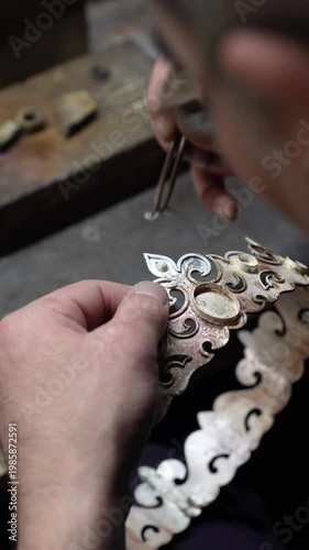 Jeweler crafting ornate silver crown with fleur-de-lis motif and detailed patterns in workshop. Traditional jewelry making process in workshop environment
