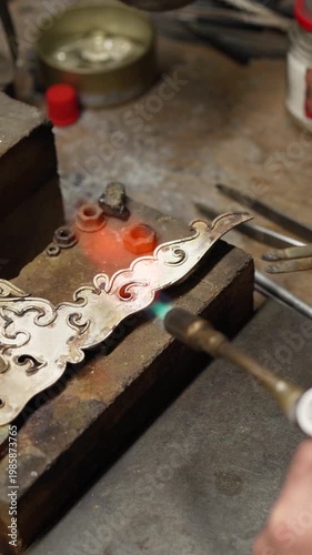 Jeweler crafting ornate silver crown with fleur-de-lis motif and detailed patterns in workshop. Traditional jewelry making process in workshop environment
