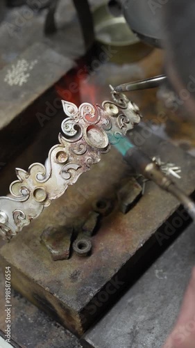 Jeweler crafting ornate silver crown with fleur-de-lis motif and detailed patterns in workshop. Traditional jewelry making process in workshop environment
