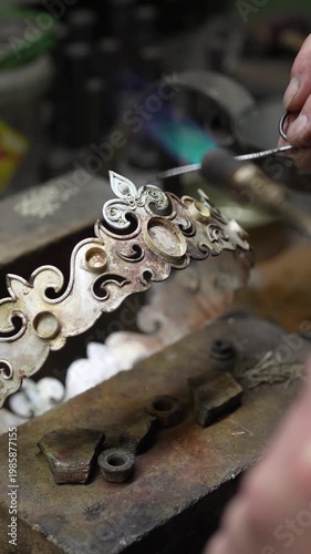 Jeweler crafting ornate silver crown with fleur-de-lis motif and detailed patterns in workshop. Traditional jewelry making process in workshop environment
