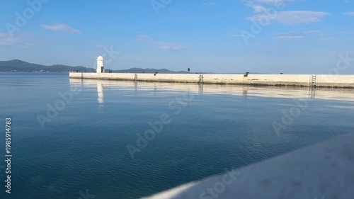 A group of people strolls along the pier while enjoying the sunlight at the coast. Calm water reflects the sky. The lighthouse stands on the pier with some boats nearby.