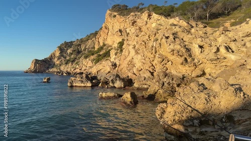 The sun sets over the rocky coastline. Waves gently hit the shore. A calm atmosphere surrounds the area. People walk nearby, enjoying the natural beauty.