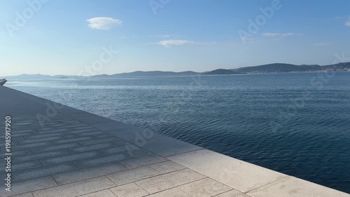 A view of clear blue water along the coast shows boats moving slowly across the sea. The sun shines bright in the sky, and distant islands can be seen.