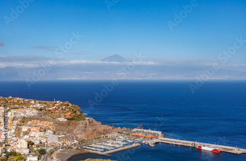 Coastal view of La Gomera with harbor and cliffs, overlooking the Atlantic Ocean, with Mount Teide on Tenerife rising above clouds in the distance under a clear blue sky