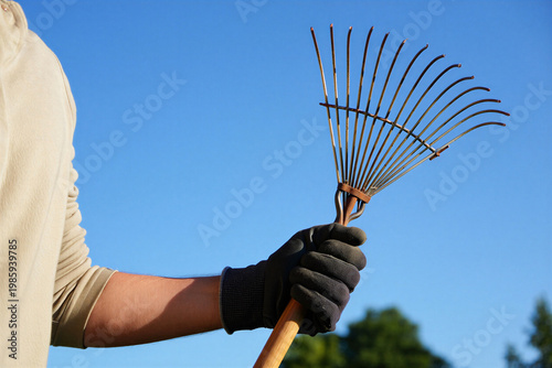 Hand holding a garden rake against a clear blue sky gardening tool green leaves