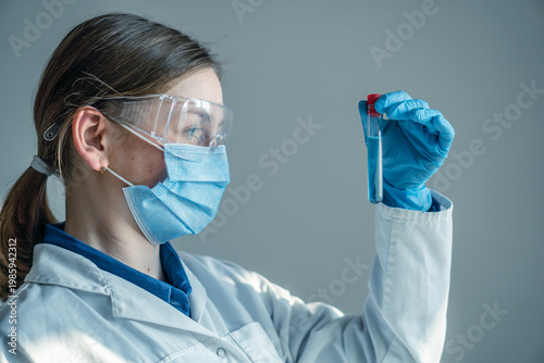 A young woman scientist wearing safety equipment holds up a test tube for analysis in a bright laboratory. The image highlights scientific research, healthcare, and laboratory safety protocols