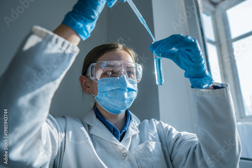 A young woman scientist wearing safety equipment carefully transfers liquid with a pipette into a test tube. The laboratory setting highlights scientific research and innovation