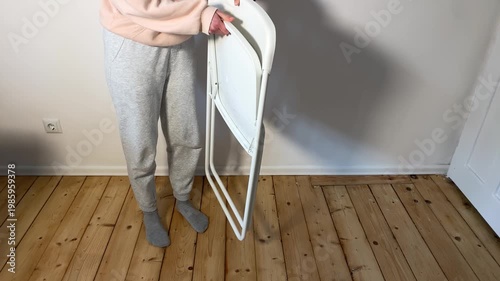 A person in casual wear unfolds a white folding chair inside a room with a wooden floor. This clip shows the setup process for temporary or event seating.