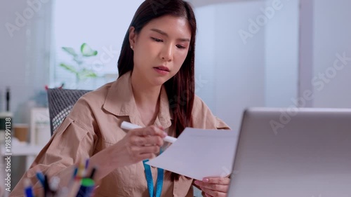 Young asian businesswoman looking stressed holding paper document working on laptop computer at corporate office desk showing serious expression feeling frustrated overwhelmed overworked tired
