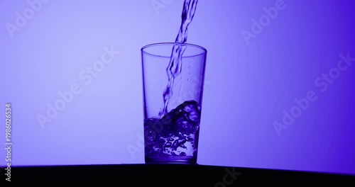 Slow-motion stream of water pouring into a clear glass, creating bubbles and splashes under vibrant blue and purple lighting with gradient background and copy space