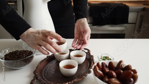 close-up of hands placing a filled white finjan cup onto a copper tray with arabic coffee, coffee beans, dates and cardamom prepared for serving