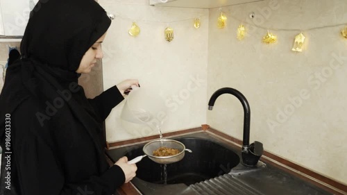muslim woman in black hijab rinses golden raisins over the sink in a home kitchen decorated with warm festive lights, documenting an ingredient-prep moment for a traditional meal