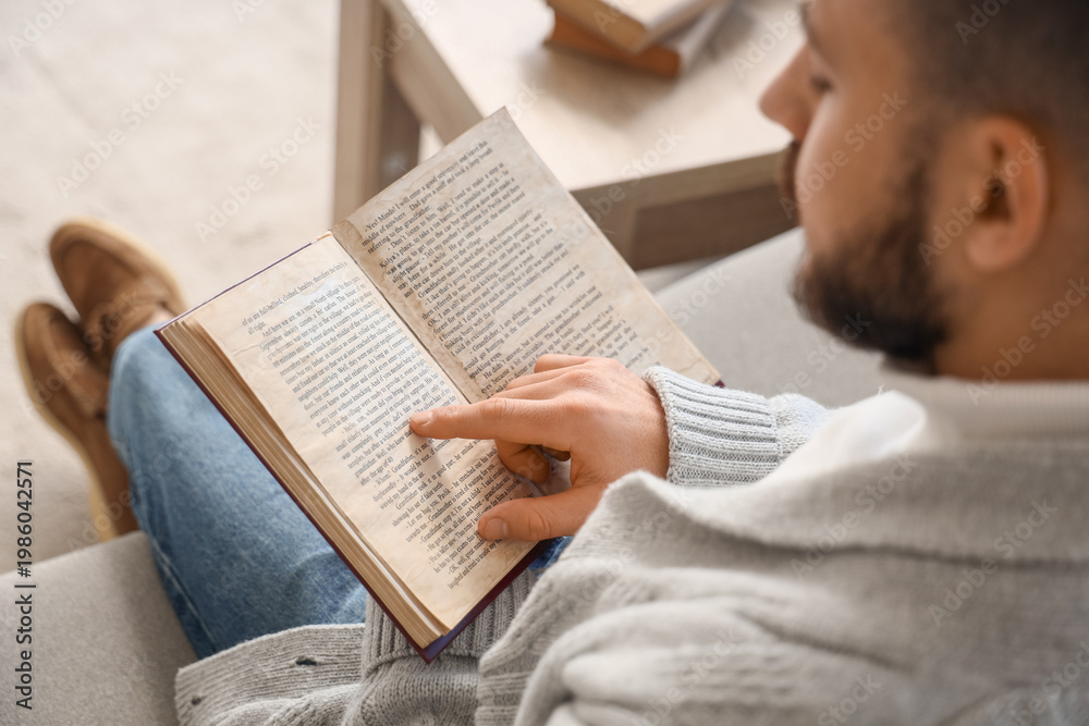 Naklejka premium Young man reading book on grey sofa at home, closeup