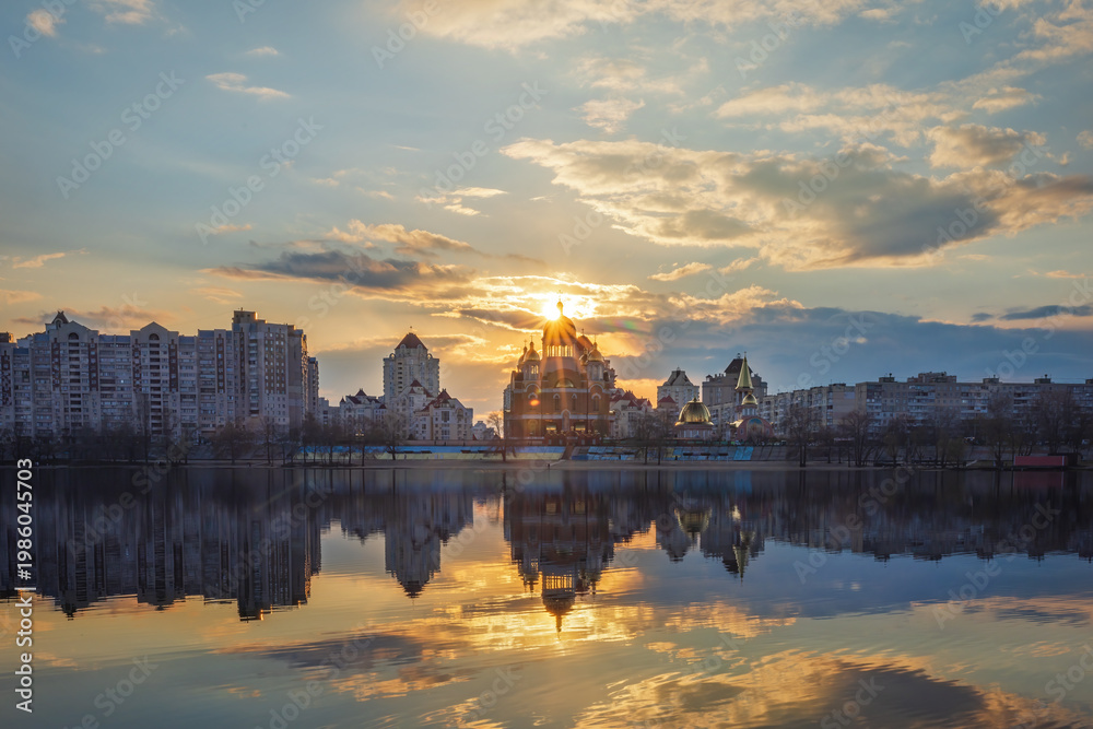 Obraz premium Golden sunset melts over Obolon embankment in Kyiv, painting the Dnipro River with shimmering reflections. Clouds drift softly as glowing domes and skyline dissolve into a calm, dreamy evening.