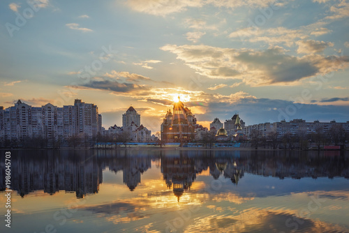 Golden sunset melts over Obolon embankment in Kyiv, painting the Dnipro River with shimmering reflections. Clouds drift softly as glowing domes and skyline dissolve into a calm, dreamy evening.