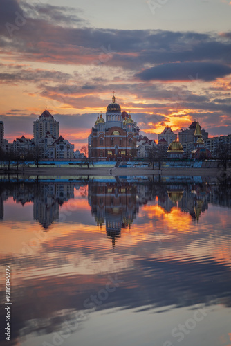 Golden sunset melts over Obolon embankment in Kyiv, painting the Dnipro River with shimmering reflections. Clouds drift softly as glowing domes and skyline dissolve into a calm, dreamy evening.