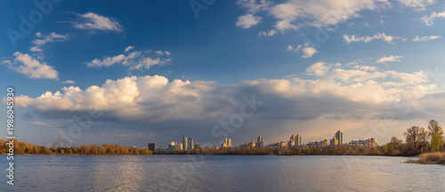 Wide panorama of Obolon embankment in Kyiv, where golden evening light touches the Dnipro River, dramatic clouds drift above the skyline, and the city glows in a serene riverside atmosphere.