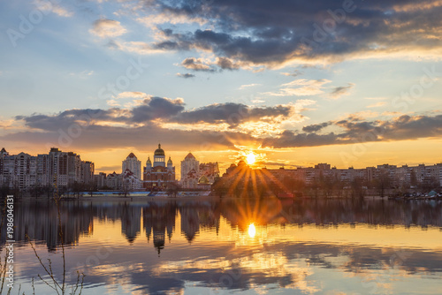 Golden sunset melts over Obolon embankment in Kyiv, painting the Dnipro River with shimmering reflections. Clouds drift softly as glowing domes and skyline dissolve into a calm, dreamy evening.