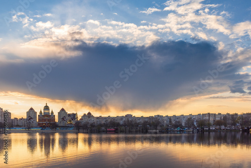 Golden sunset melts over Obolon embankment in Kyiv, painting the Dnipro River with shimmering reflections. Clouds drift softly as glowing domes and skyline dissolve into a calm, dreamy evening.