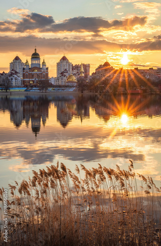 Golden sunset melts over Obolon embankment in Kyiv, painting the Dnipro River with shimmering reflections. Clouds drift softly as glowing domes and skyline dissolve into a calm, dreamy evening.