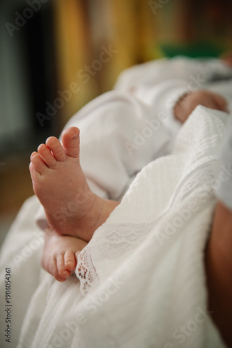 Close-up of a baby's tiny foot during a baptism ceremony. The infant is wrapped in a white lace garment, creating a peaceful and sacred atmosphere in the church.