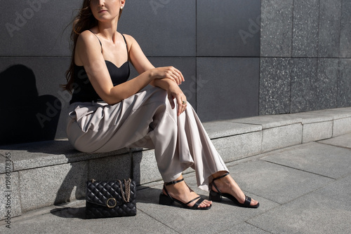 Woman in black heeled sandals, beige palazzo pants, black tank top and small black bag sitting outside on city street. Trendy elegant casual outfit. Street fashion. Women's legs, without a face.