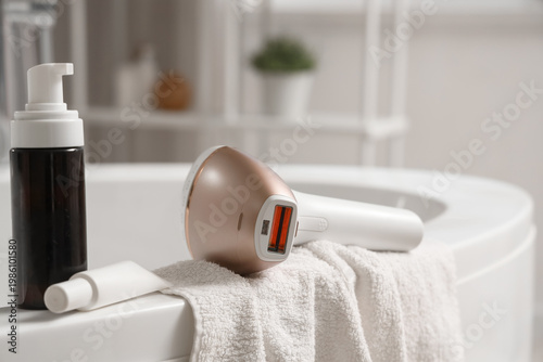 Foto Modern photoepilator with towel and cosmetic products on bathtub in bathroom