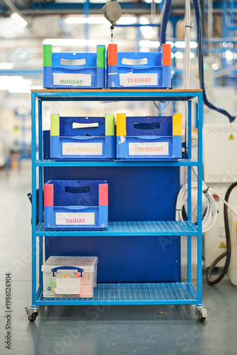 Organised production trolley with labelled plastic bins for each weekday in a bright factory setting, vivid blue structure with colourful labels creating a structured, efficient and orderly atmosphere