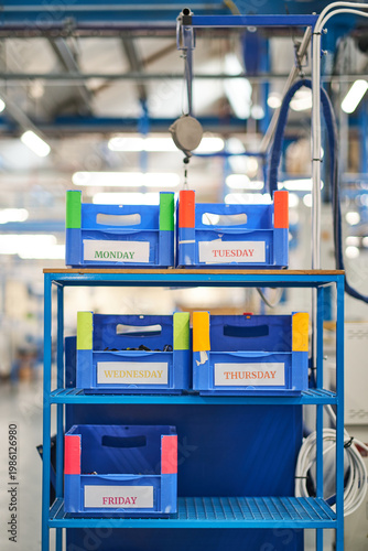 Organised production trolley with labelled plastic bins for each weekday in a bright factory setting, vivid blue structure with colourful labels creating a structured, efficient and orderly atmosphere