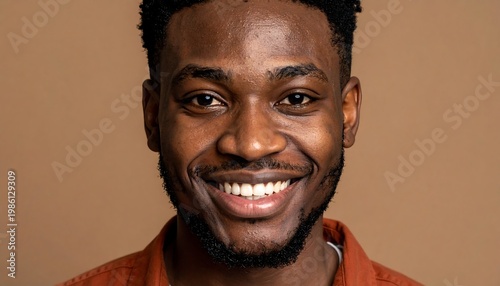 Smiling close-up of a young, dark-skinned man with a brown shirt against a neutral tan background