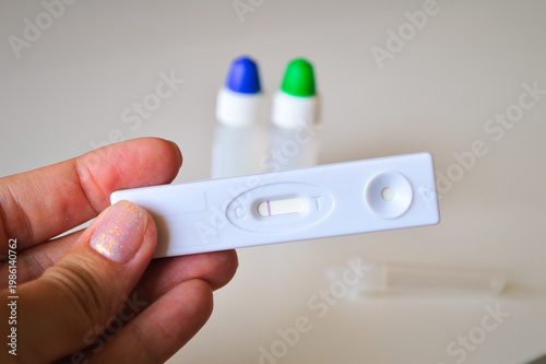 Close-up of a female hand holding a negative rapid antigen diagnostic test cassette with medical buffer solution bottles in a bright blurred background