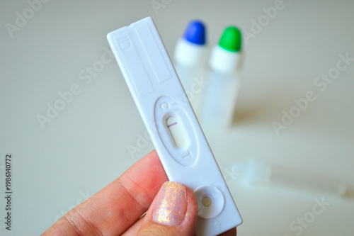 A hand with glittery nails holding a white diagnostic rapid test cassette showing a single line result in a bright medical laboratory environment.
