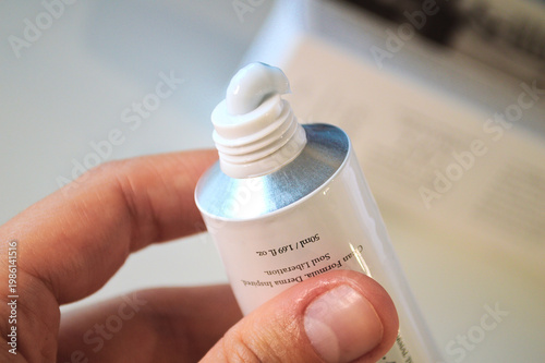 Close-up view of a person's hand gently squeezing a light blue moisturizing face cream from a white aluminum cosmetic tube against a bright background for daily skincare routine.