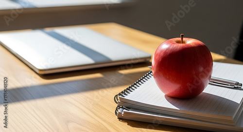 Fresh red apple on spiral notebook with pen and laptop on wooden office desk in bright sunlight, healthy snack at work and education motivation concept, realistic still life photography.