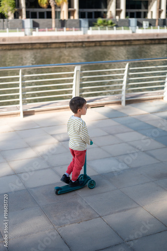 Young Boy Riding A Kick Scooter Along The Waterfront Promenade.