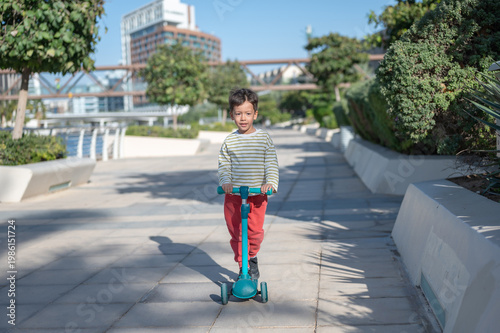 A cheerful five-year-old boy rides a turquoise scooter along a paved pathway in a contemporary urban waterfront promenade.