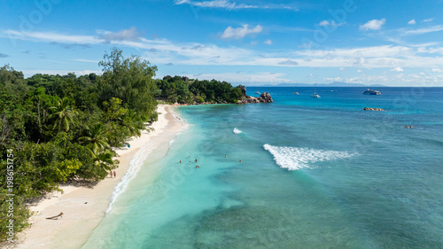 Turquoise water washing over white sandy beach bordered by dense green vegetation. La Digue, Seychelles.