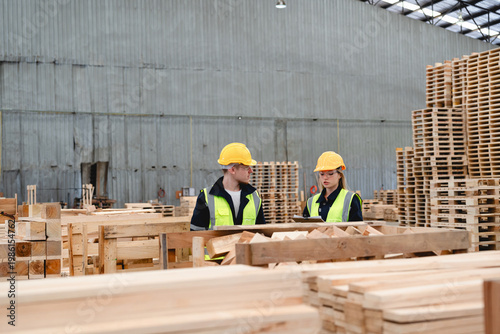 Photos Two Workers in Yellow Hard Hats and Neon Vests Inspect Wooden Pallets During Warehouse Quality Control Assessment in a Large, Dimly Lit Industrial Facility