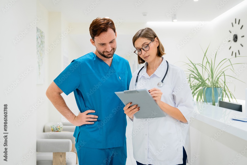 Fototapeta premium Male surgeon and young female doctor looking at clipboard with patient information in hospital corridor, medical teamwork, professional communication, healthcare staff collaboration and clinic service