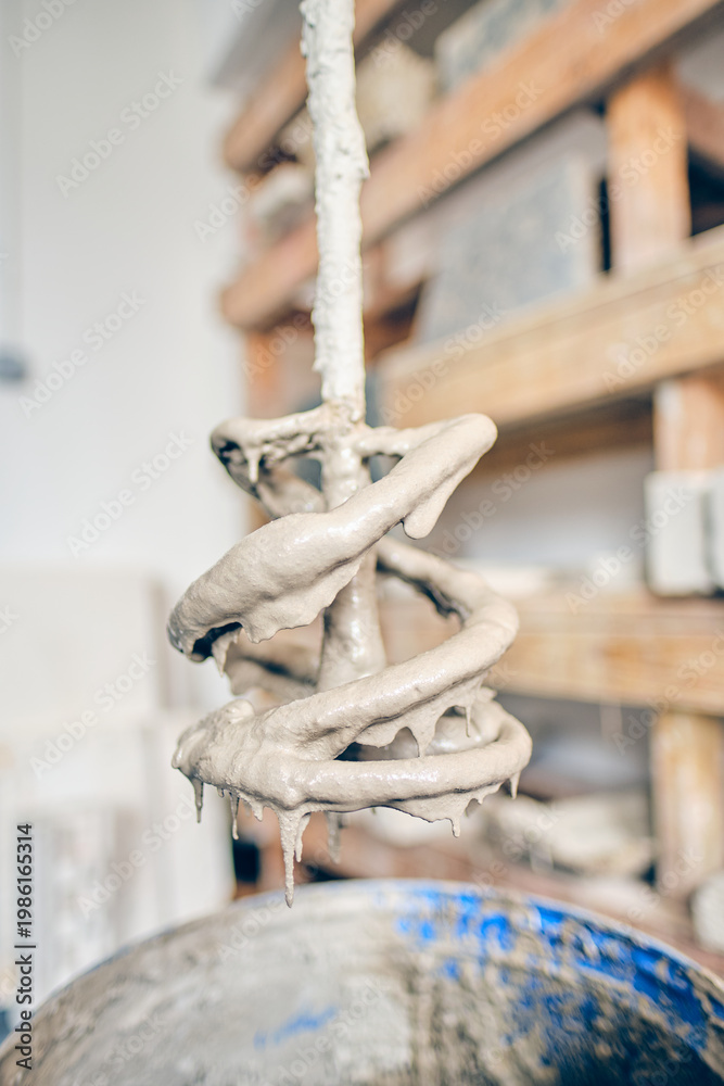 Fototapeta premium Close up of a drill mixer coated in raw gritty wet plaster or cement suspended above a mixing bucket, cool grey slurry dripping from the spiral blade against a soft workshop background,