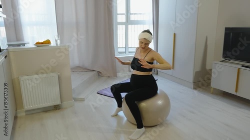 Prenatal exercises and fitness at home. Pregnant woman sits on fitball, moves her arms out to sides, does exercises in her living room. Preparing for childbirth in the seventh month of pregnancy.