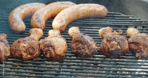 Chicken drumsticks and sausage links cook over a smoky charcoal grill