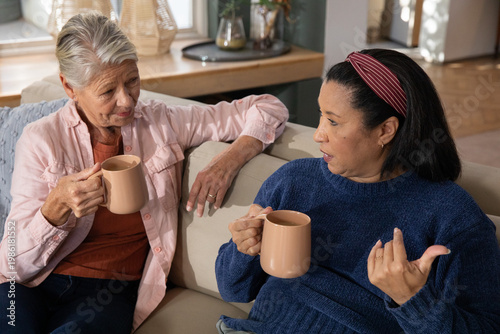 Diverse senior female friends sitting on beige sofa holding matching tan ceramic mugs and chatting