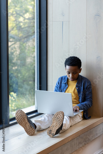 African American boy using laptop on window bench wearing denim jacket, sneaker soles, copy space