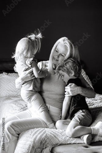 Happy mother cuddling and playing with two young children on a bed in a cozy bedroom.