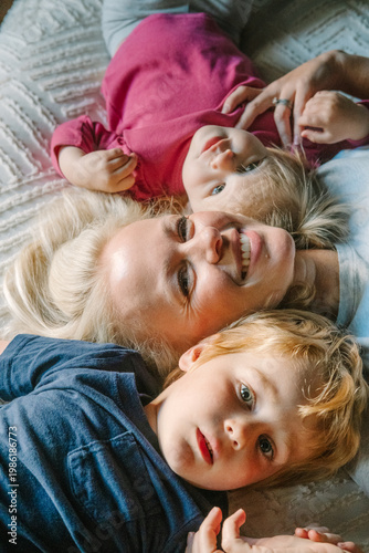 Smiling mother and two young children lying together on a bed, sharing a joyful family moment.