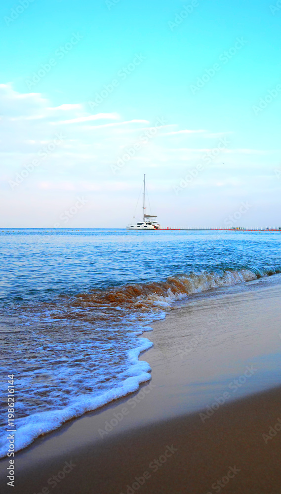 Fototapeta premium Sea waves at sandy beach and sailing catamaran moored at pier in background. Catamaran is used for cruises and charters to Asian resorts. Sailing catamaran anchored in calm waters at shore.
