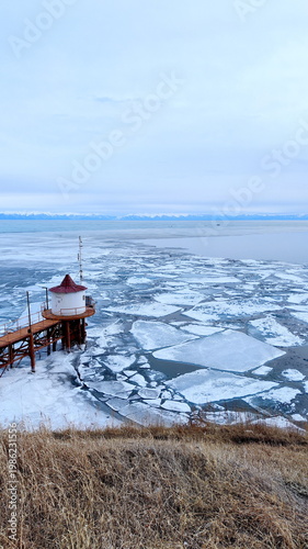 View from Shore to Frozen Lake Baikal with Old Metal Bridge and Building.